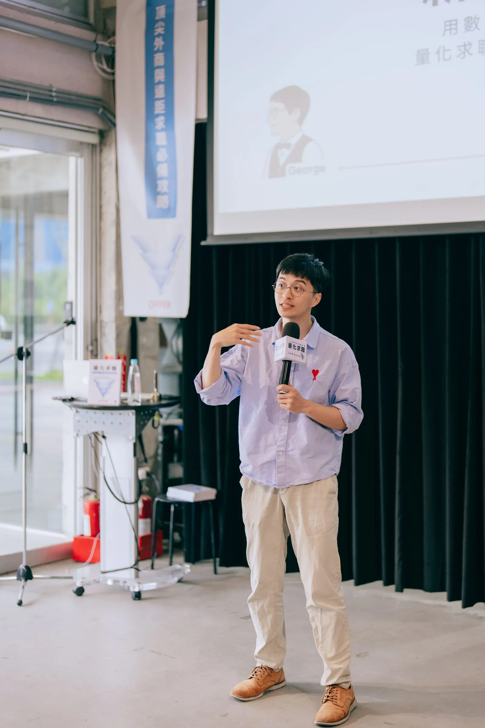 企業引導師喬拿著麥克風分享，背後是投影幕與直立旗